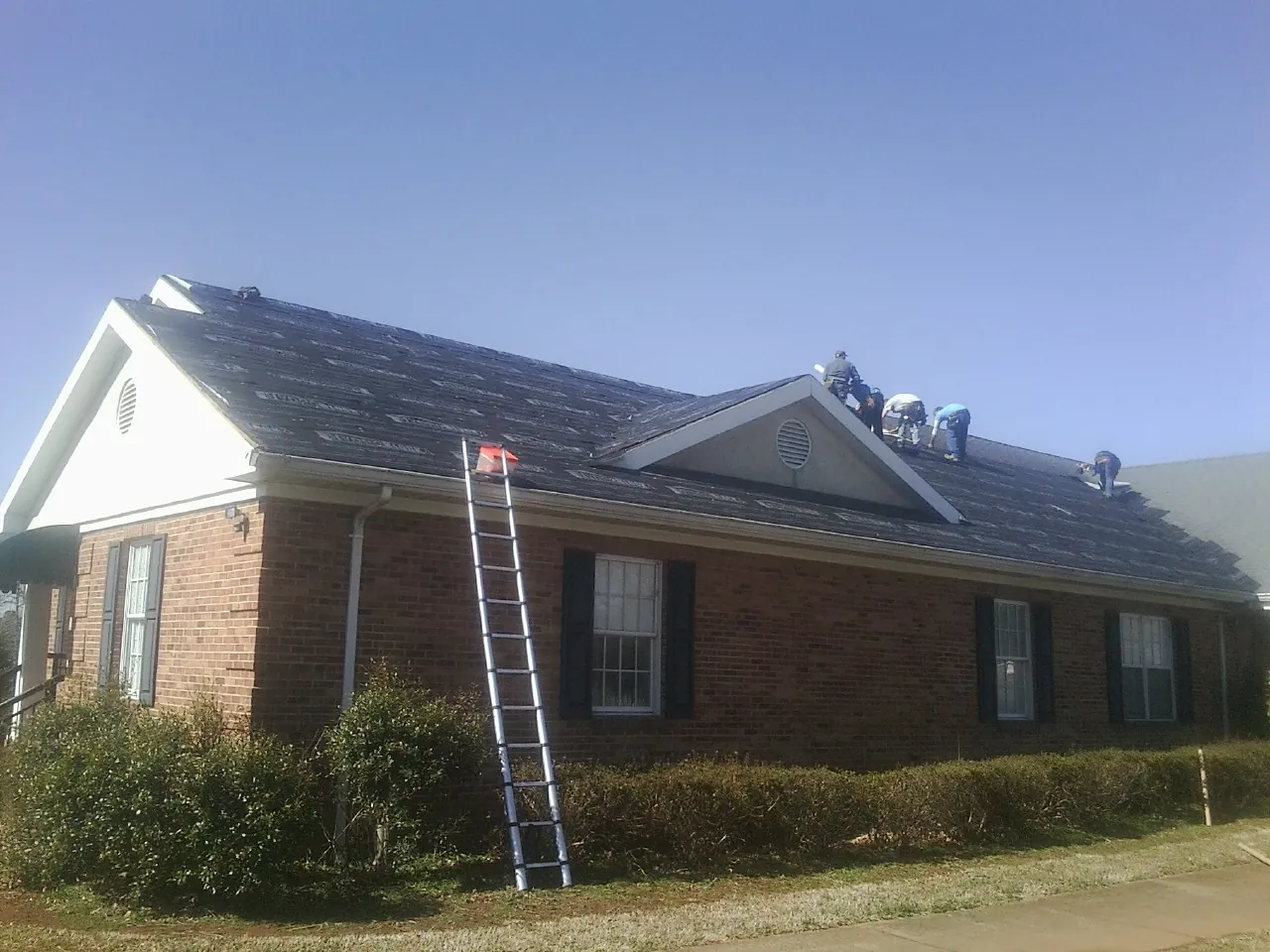 Crew tearing shingles off a brick ranch roof with a ladder set — Emergency Roof Repair work by our crew in Big Arm